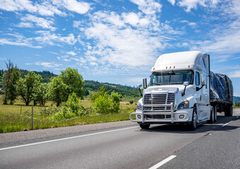 Elegant white big rig semi truck with chrome grille guard transporting tarp cargo on flat bed semi trailer driving on the straight road at sunny day with cloud sky