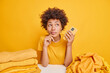 © WHstudio Leushin N - Pensive Afro American woman has dreamy expression holds modern mobile phone sits at table with piles of clothes isolated over yellow background busy folding laundry. Clothing and housekeeping