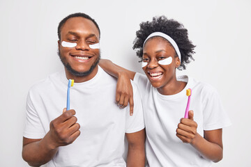 Happy dark skinned woman and man have fun undergo beauty and hygiene procedures hold toothbrushes going to clean teeth dressed in casual t shirts isolated over white background. Daily routine