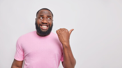 Wall Mural - That way. Positive dark skinned man with white teeth points thumb away on blank space shows place for logo or product placement dressed in casual pink t shirt isolated over white background.