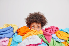 Wondered Curly Haired Ethnic Woman Focused Above Surrounded By Multicolored Laundry Cluttered With Clothes Collects Clothing For Recycling Isolated Over White Background. Organize Your Closet