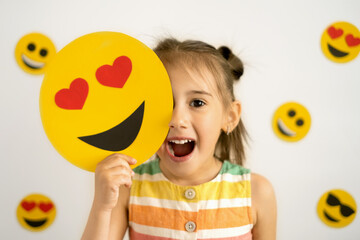 Sticker - A little girl, smiling with her mouth open, holds in her hands a cardboard love smile with hearts instead of eyes, covering part of her face with them. World emoji day. Anthropomorphic smile Face.