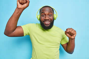 Wall Mural - Joyful bearded Afro American man moves with rhythm of music wears wireless headphones on ears dressed in green t shirt has fun isolated over blue background. People entertainment and hobby concept