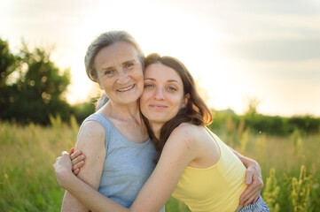 Senior mother with gray hair with her adult daughter looking at the camera in the garden and hugging each other during sunny day outdoors