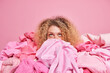 Young woman with curly hair focused above puts things in order surrounded by stack of clothes collects garments isolated over pink background. Horizontal monochrome shot. Clothing revision concept