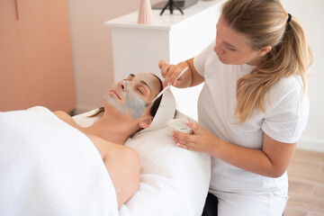 Cleaning and moisturise nourish skin of face. Cosmetologist put grey mask on face by brush. Beautiful woman receiving cavitation facial peeling. Treatment skin care in cosmetology beauty spa salon.