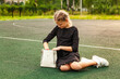 A blonde schoolgirl in a black dress sits on the green floor of the sports ground. Daytime, warm day, white handbag. Swarms in a bag