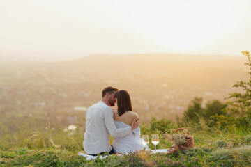 Rear view of hugging couple resting on the mountain at summer picnic at beautiful sunset.