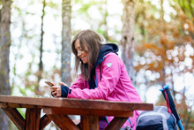 Hiker Girl Resting On A Bench In The Forest. Backpacker With Pink Jacket Holding Cell Phone.