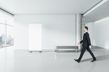 Aufkleber - Businessman walking in modern gallery interior with empty poster on concrete wall and window with city view. Mock up.