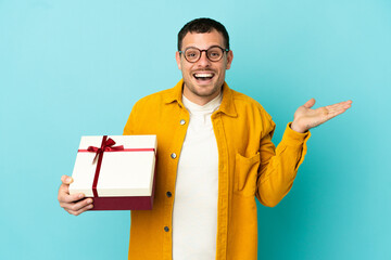 Brazilian man holding a gift over isolated blue background with shocked facial expression