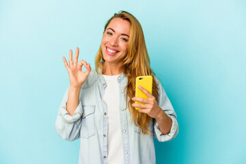 Caucasian woman holding mobile phone isolated on blue background cheerful and confident showing ok gesture.