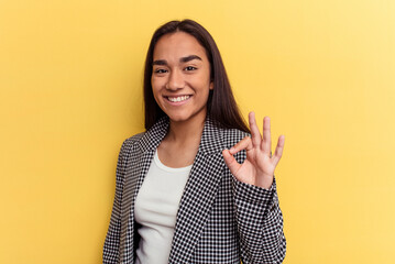 Wall Mural - Young mixed race woman isolated on yellow background winks an eye and holds an okay gesture with hand.