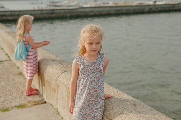 two little girl with long white hair in a dress looks into the distance on the pier of the sea in summer