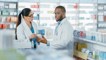 Wall Mural - Pharmacy: Black Male and Caucasian Female Pharmacists Use Digital Tablet Computer Talk about Medicine, Drugs, Vitamins, Supplements, Vaccine, Health Care Products. Medical Professionals in Drugstore