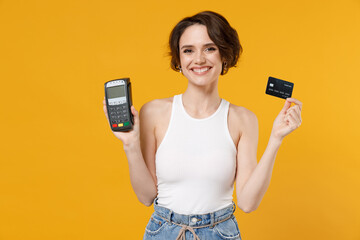 Young happy smiling rich woman 20s wearing white tank top shirt holding wireless modern bank payment terminal to process and acquire credit card payments isolated on yellow color background studio