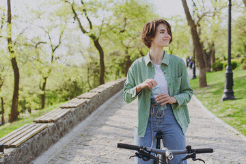 Young woman 20s in green jacket jeans riding bicycle bike in city spring park outdoors, look aside drink clear fresh pure water from transparent glass of water People active healthy lifestyle concept