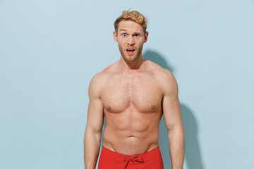 Handsome young sexy shock confused sporty man in red shorts swimsuit relax near hotel pool looking camera with open mouth isolated on pastel blue background Summer vacation sea rest sun tan concept