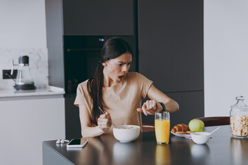 Young disappointed housewife woman 20s in t-shirt eating late breakfast in morning look at smart watch check time hurrying rush cooking food in light kitchen at home Healthy diet lifestyle concept