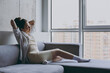 Full length side view young relaxed caucasian woman 20s in casual white clothes sit on soft grey sofa near window hold hands behind neck yawning indoors apartment Resting leisure staying home concept