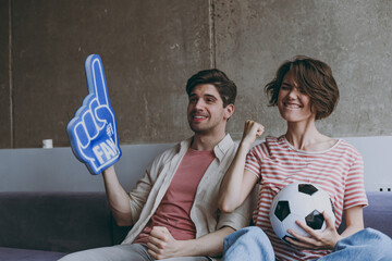 Wall Mural - Winner couple friends woman man football fans 20s in casual clothes foam glove finger up support favorite team clench fist with soccer ball scream indoors at home watch tv People sport family concept.