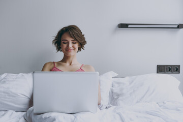Calm smiling young woman in pajamas lies in bed wrap covered under blanket duvet on pillow hold use work on laprtop pc computer look aside rest relax indoors at home Good mood morning bedtime concept