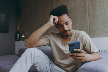 Sticker - Pensive thoughtful sad young african american man wearing beige t-shirt sit on grey sofa indoors apartment using mobile cell phone prop up forehead think on weekends stay home. Tattoo translate fun.