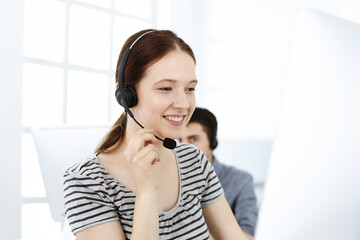 Casual dressed young woman using headset and computer while talking with customers online. Group of operators at work. Call center, business concept