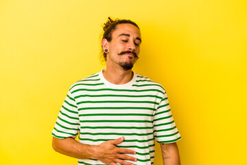 Young caucasian man with long hair isolated on yellow background touches tummy, smiles gently, eating and satisfaction concept.