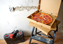 Pizza For Lunch On The Stairs. Take A Break From Work. Black Ladder, Hard Hat, Ear Protectors, Protective Gloves And Tool Case Against The Background Of A Concrete Wall. Renovation Concept.