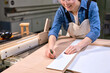 Young cropped caucasian female carpenter in blue shirt using pencil drawing line with a measurement tool on wooden plank on table in workshop, enjoy carpentry, woodworking