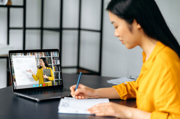 Wall Mural - Clever asian female student gains knowledge watching lesson online and studying distantly. Young woman takes notes during webinar, listening lecture from female teacher on video call. Online education