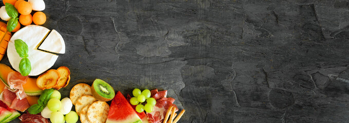  Summer fruit theme charcuterie corner border against a dark slate background. Selection of fruits, cheese and meat appetizers. Copy space.