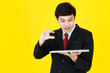 Horizontal portrait shot of young adult Asian man in a black long sleeve suit holding a closed laptop while trying to touch something in empty space  in air isolated with yellow background in studio