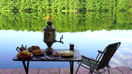 Wall Mural - Vintage metal tea samovar with white smoke and food on the table near the calm water lake in green forest at morning, Ukraine. Nature food concept