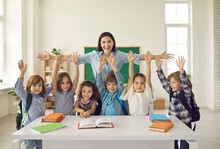 Happy Teacher With Arms Widely Spread To Hug Cheerful Children Raising Hands. Positive Portrait In Classroom. Educator Showing Love, Adoration And Care For Pupils. Children And Tutor Looking At Camera