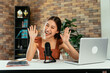 © twinsterphoto - Portrait of cheerful young Asian female blogger using mic and laptop with digital tablet to interview guest during live stream at home