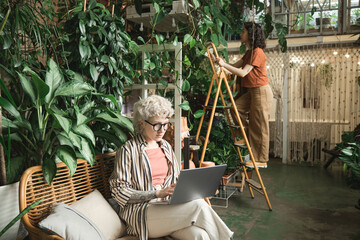 Wall Mural - Woman working on laptop in the flower shop with her colleague caring about plants in the background