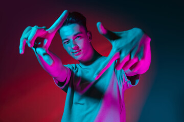 Gesturing. Portrait of Caucasian young man posing isolated on dark red studio background in neon pink light. Concept of human emotions, facial expression.