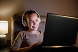 Fototapeta Tulipany - Happy young woman in headphones working or studying at night on sofa using laptop. Teenager girl resting studing watching movie on laptop. internet surfing