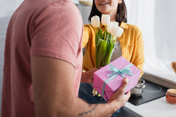 partial view of young man presenting bouquet of flowers and gift box to girlfriend in kitchen