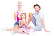 © Mediaphotos - Horizontal studio portrait of family with cute daughter wearing summer outfits sitting on floor drinking juice and eating ice cream, white background