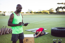 African american male fitness instructor using tablet, with colleague exercising outdoors