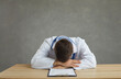 © Studio Romantic - Tired and exhausted male doctor sleeps sitting at a table after a hard day's work. Man put his head on the table sitting on a gray concrete background. Concept of stress and fatigue of medical staff.