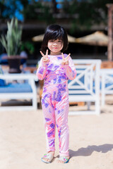 vertical imaged. Happy Asian cute girl standing and posting take a picture. Child looking at camera. Sweet smiling. At the beach. Summer time. Children wearing swimsuit. Baby aged 4-5 years old.