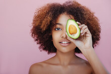 Portrait Of A Beautiful Young Dark-skinned Woman With Curly Hair Covers One Eye With Half An Avocado Looks At The Camera And Smiles On A Pink Background.