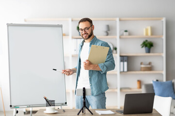 Poster - Young male teacher pointing at empty blackboard during web lesson, using smartphone and laptop at home, space for design