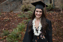 A Beautiful Young Woman In Cap And Gown With Flower Lei Smiling At Her College Graduation.