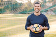 © Austockphoto - Close up shot of a smiling young man standing on the field , holding a soccer ball with a net behind