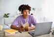 © Prostock-studio - Intelligent black teen student in headphones studying online in front of laptop, holding notebook at home